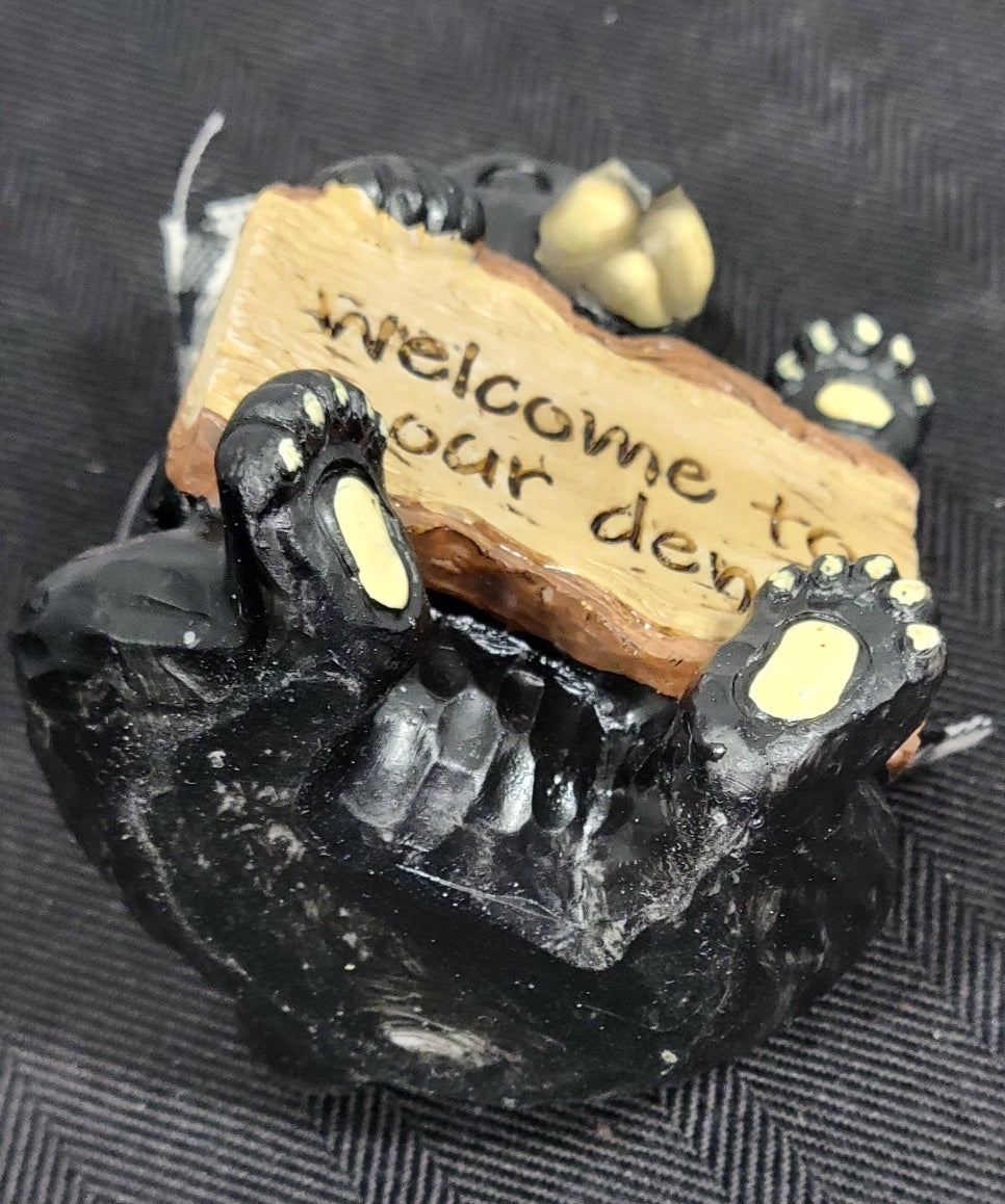 Small black bear figurine holding a wooden sign with text on a textured surface